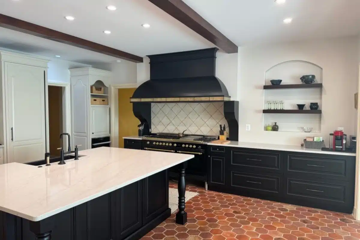 Open concept kitchen remodeling in Baltimore, MD featuring a spacious layout with a large island, black cabinetry, custom range hood, exposed ceiling beams, and warm terracotta flooring.