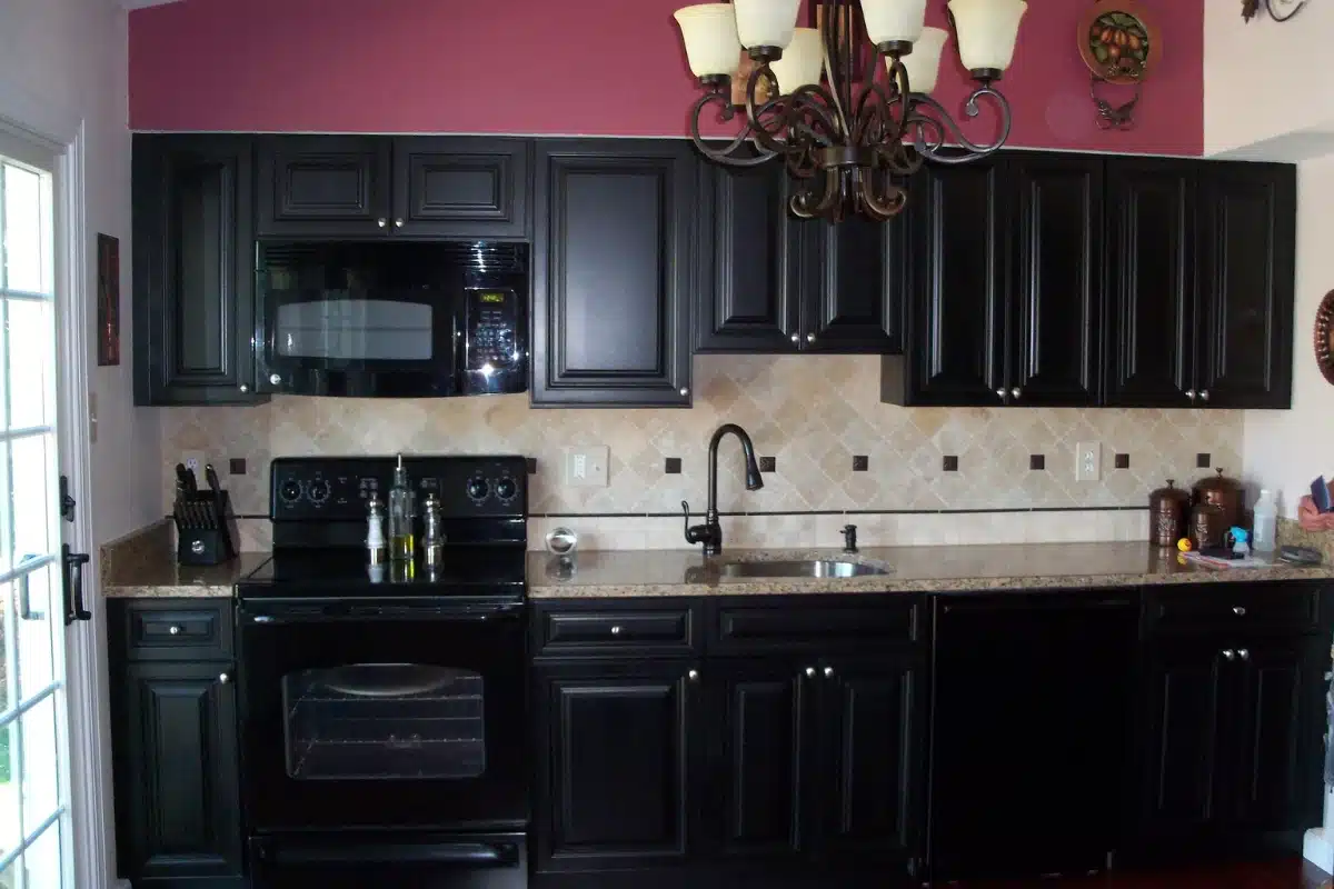 Sink installation in Baltimore, MD featured in a traditional kitchen with black cabinetry, tiled backsplash, granite countertops, and a central under-mount sink beneath decorative lighting.