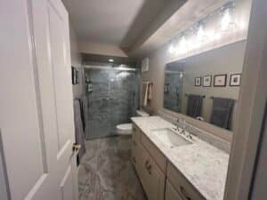 A modern bathroom renovation in Essex, MD featuring a large walk-in glass shower with marble-style wall and floor tiles, a wide white vanity with a speckled quartz countertop, and industrial-style vanity lighting.