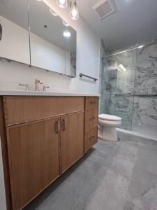 A modern master bathroom renovation in Pikesville, MD featuring a custom wood-grain vanity, white quartz countertop, large-format grey floor tiles, and a walk-in glass shower with marble-patterned wall tile.