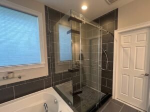 A luxury master bathroom renovation in Silver Spring, MD, featuring a frameless glass corner shower with charcoal gray horizontal tile, a black penny-tile shower floor, and a deep soaking tub.
