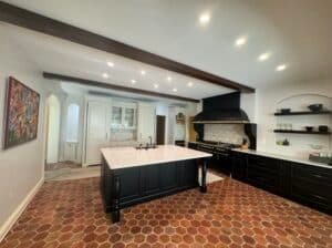 A spacious luxury kitchen renovation in Columbia, MD featuring a large black island, white perimeter cabinetry, dark wood ceiling beams, a black professional range with a custom hood, and hexagonal terracotta tile flooring.