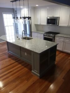 A modern kitchen renovation in Dundalk, MD featuring a large grey center island with a speckled granite countertop, white shaker perimeter cabinets, stainless steel appliances, and a linear 5-light pendant fixture over the sink.
