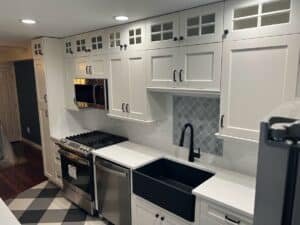 A modern kitchen renovation in Laurel, MD featuring white shaker cabinets with glass-front uppers, a black farmhouse apron sink, a gray arabesque tile backsplash accent, and bold diamond-patterned floor tile.